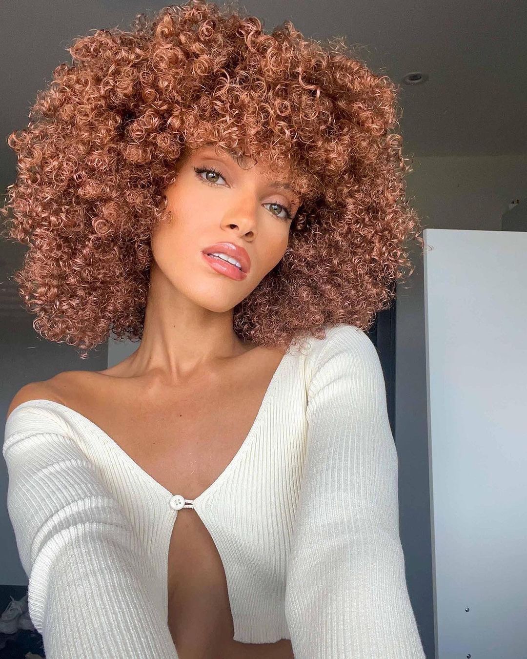 a woman with mid length tightly coiled curls and pink-copper highlights taking a selfie